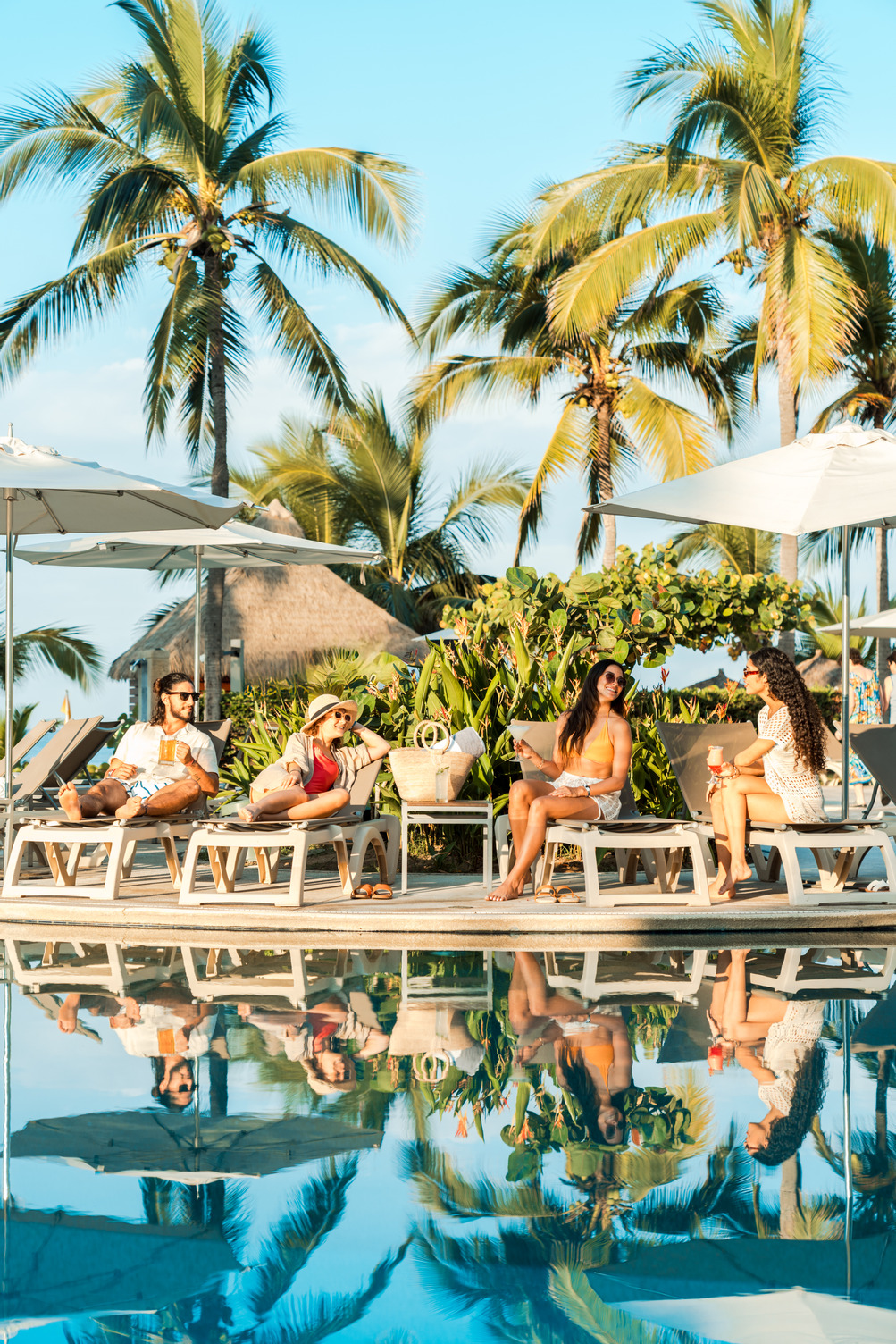 Group of people relaxing on lounge chairs by a pool with palm trees and clear blue sky in the background.