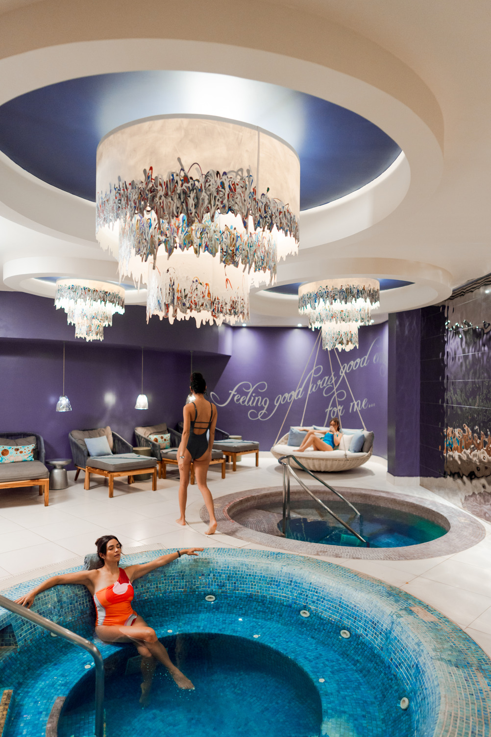 Women relaxing in a luxurious spa with elegant chandeliers, vibrant purple walls, and stone-tiled jacuzzi pools.