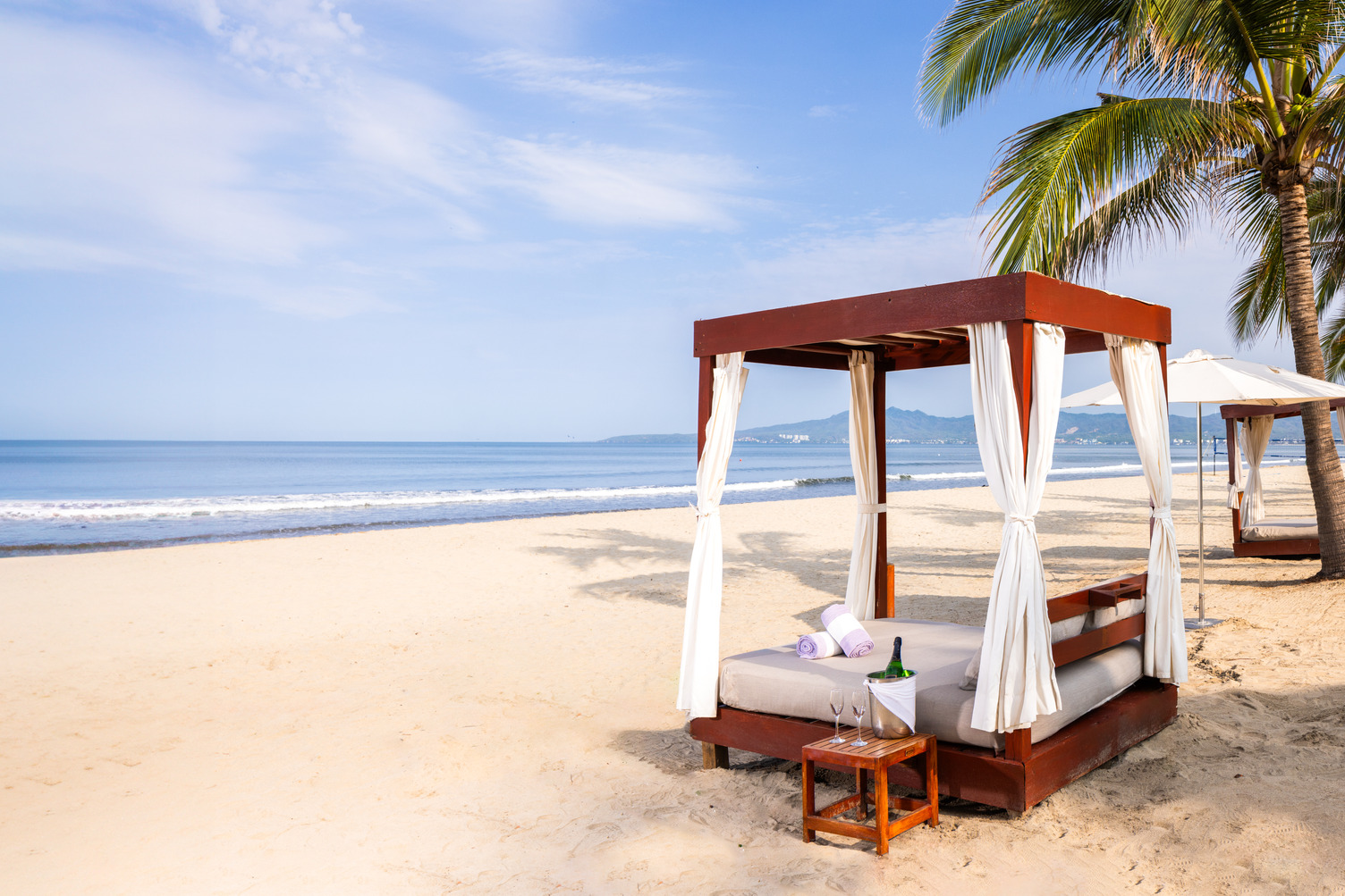 Balinese bed on a sandy beach by the ocean, sunny day, palm trees, beach chairs, and mountains in the distance.