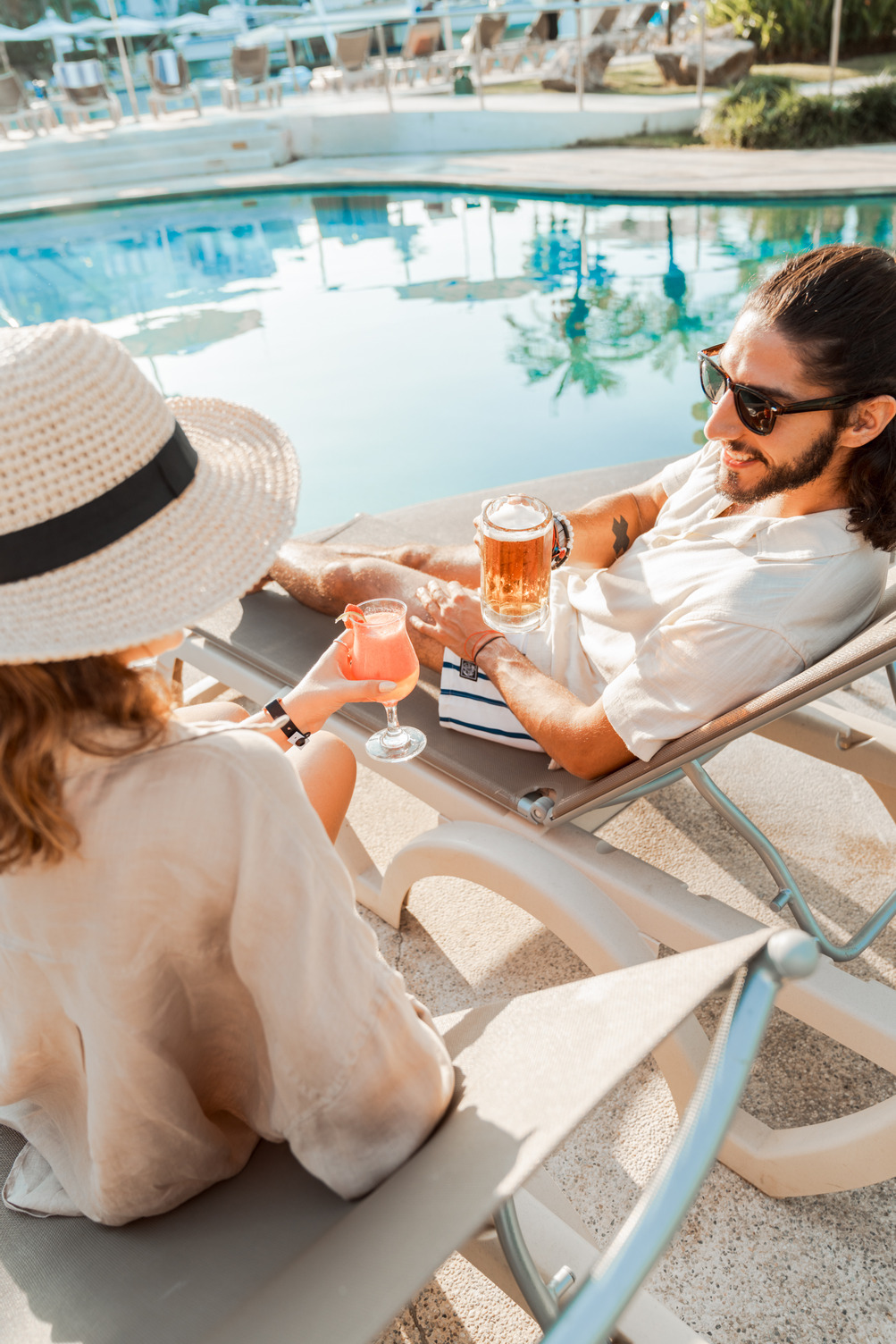 Pareja relajándose junto a la Alberca 3 con bebidas; hombre con gafas de sol sosteniendo cerveza, mujer con sombrero de sol y