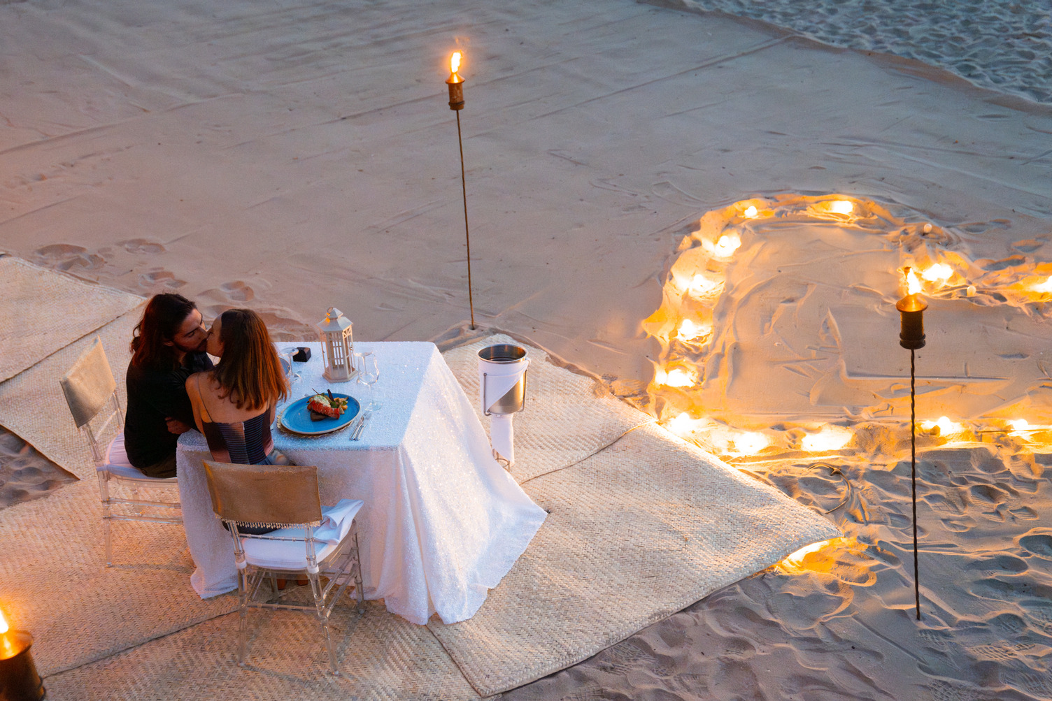 Pareja cenando en una mesa iluminada por velas en una playa de arena, rodeada de una forma de corazón hecha de luces.