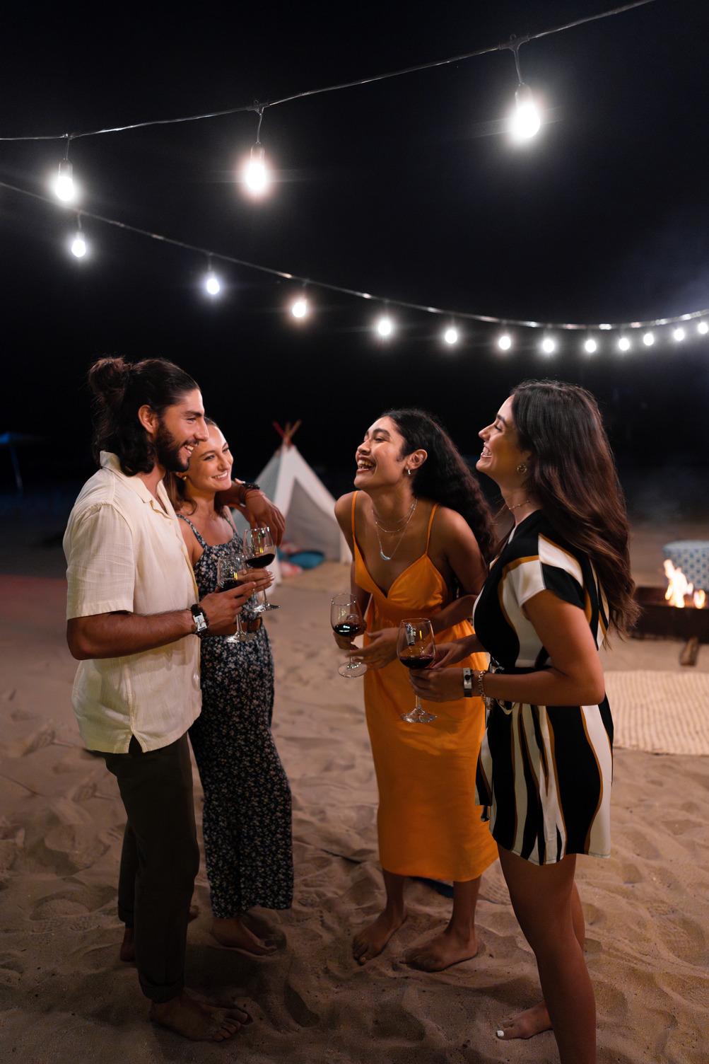 Group of four people laughing and holding drinks near a beach bonfire at night, with string lights overhead.