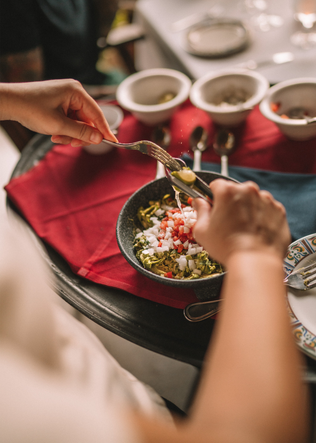 Person preparing guacamole in a bowl with diced tomatoes and onions, surrounded by condiments on a red cloth.