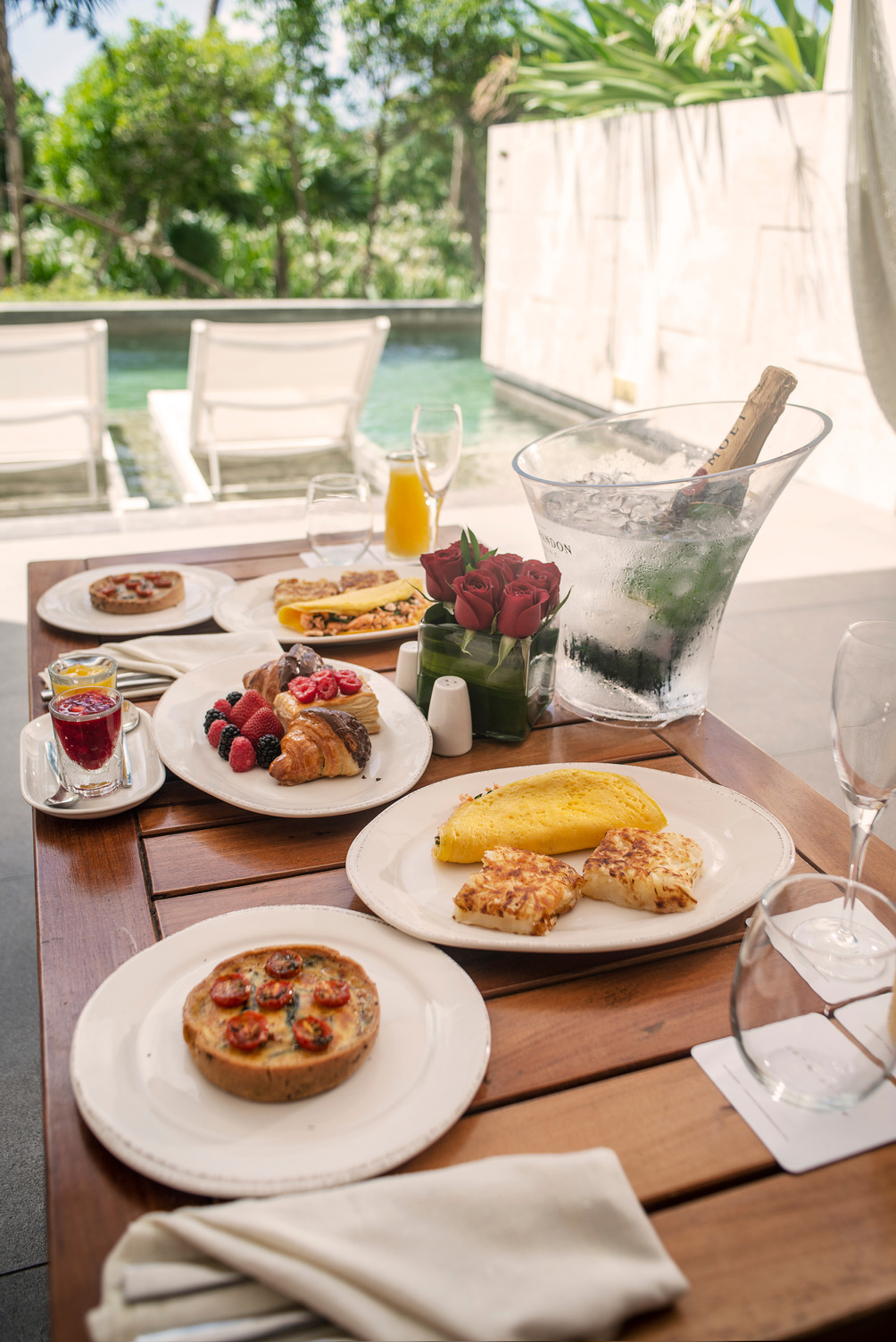 Desayuno servido en una mesa de madera con panecillos, omelette, fruta, y champaña, con vista a una alberca con camastros.