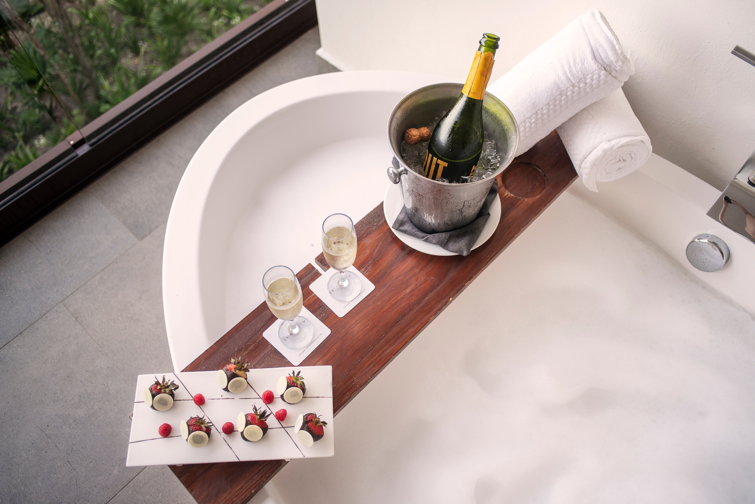 Botella de champagne en cubeta de hielo, dos copas y fresas en una bandeja junto a una bañera con burbujas.