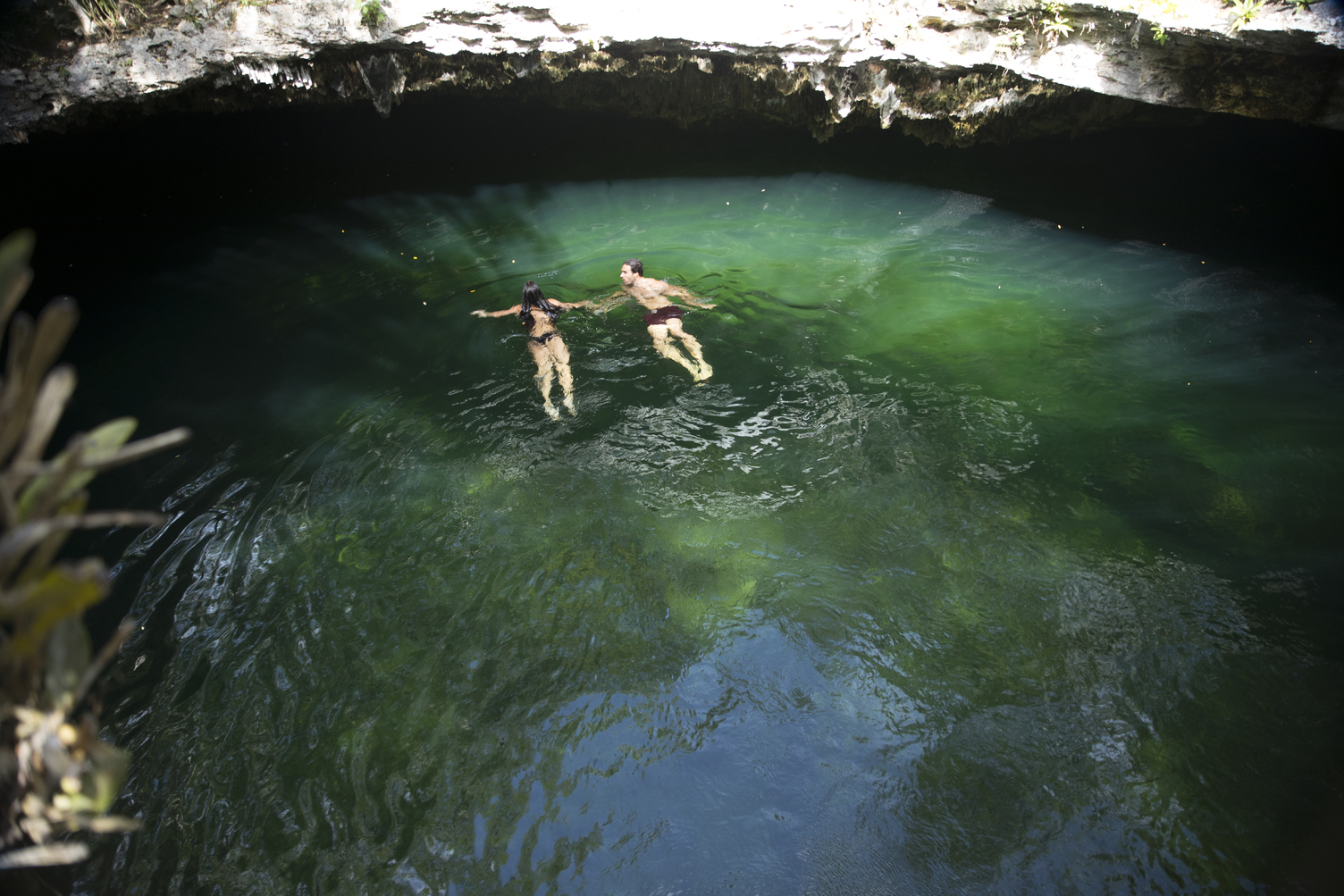 Two people swimming in a natural cenote with clear green water and rocky surroundings.