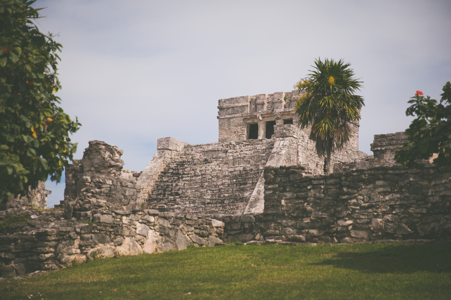 Muyil Archeological Ruins with ancient stone structures, tropical plants, and a partly cloudy sky in the background.