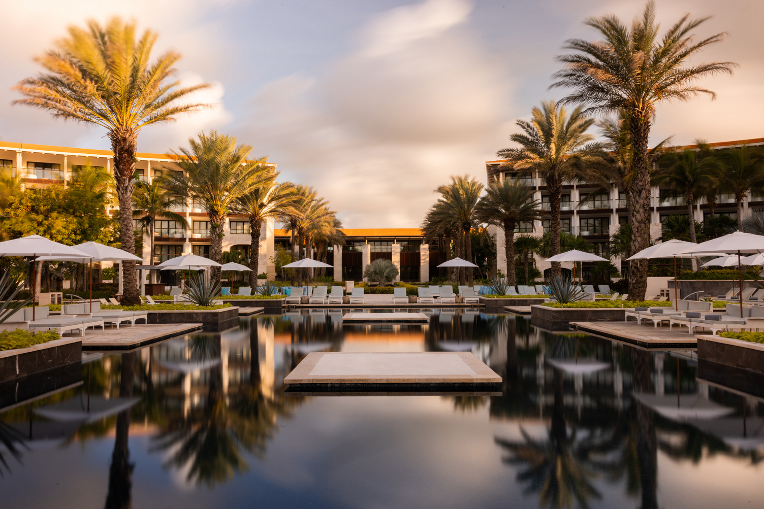 Luxury resort pool surrounded by palm trees, lounge chairs, and modern hotel buildings at sunset.