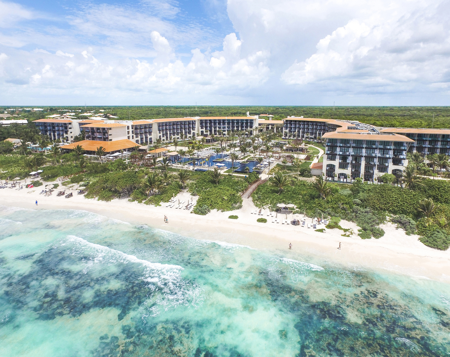 Vista aérea de un lujoso resort frente a la playa con palmeras, agua turquesa clara y playa de arena blanca en Riviera Maya.